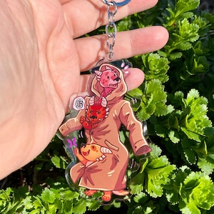 May include: A brown, red, and yellow cartoon character keychain with a pink and red dragon character holding a yellow dragon character. The keychain is made of clear acrylic.