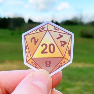 May include: A yellow and brown 20-sided die sticker with the numbers 2, 4, 6, 8, 10, 12, 14, 16, 18, 20 printed on the faces.