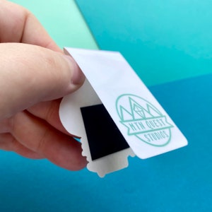 K&ouml;nnte beinhalten: Ein wei&szlig;er Aufkleber mit einer schwarzen magnetischen R&uuml;ckseite. Der Aufkleber zeigt ein gr&uuml;nes Logo mit einer Bergkette und einem Diamanten, mit dem Text "MTN QUEST STUDIOS".