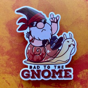 May include: A sticker featuring a cartoon gnome wearing sunglasses and a red bandana, riding a snail with flames on its shell. The text "Bad to the Gnome" is printed below the gnome.