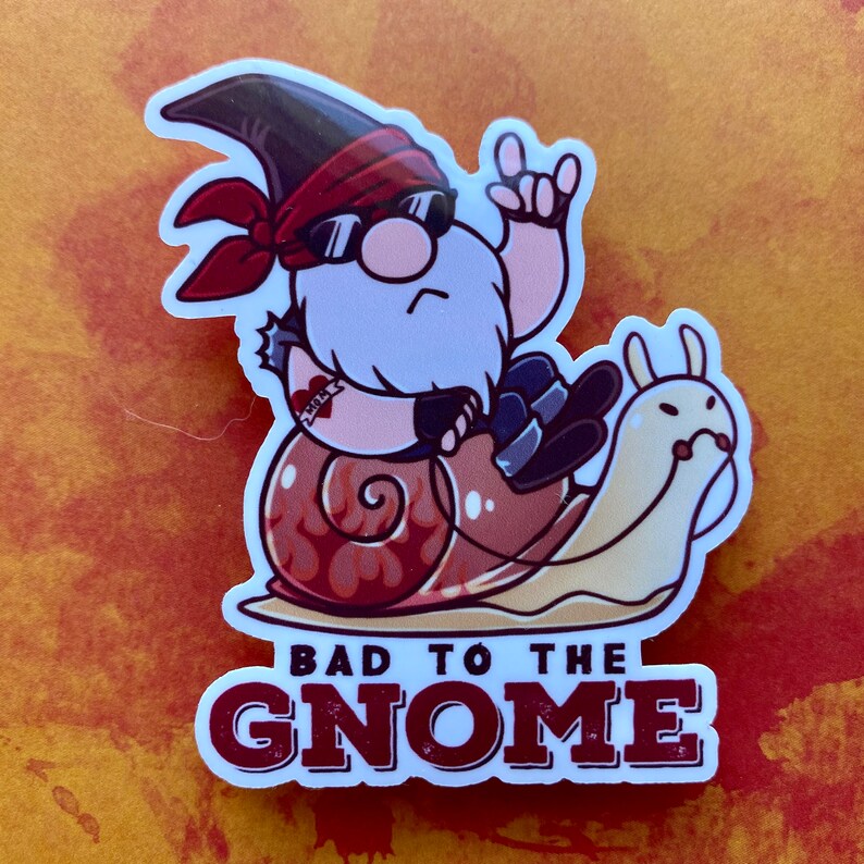 May include: A sticker featuring a cartoon gnome wearing sunglasses and a red bandana, riding a snail with flames on its shell. The text "Bad to the Gnome" is printed below the gnome.