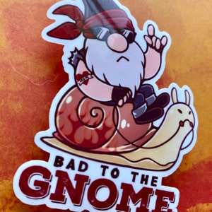 May include: A sticker featuring a cartoon gnome wearing sunglasses and a bandana, riding a snail with flames on its shell. The text "BAD TO THE GNOME" is printed below the gnome.