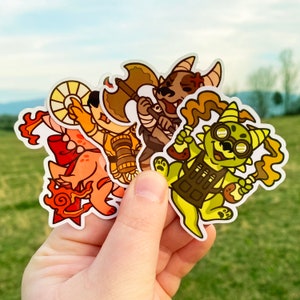May include: Three colorful cartoon character stickers. The first sticker is a red and orange creature with a large, round head and a long tail. The second sticker is a green and yellow creature with a large, round head and a long tail. The third sticker is a brown and orange creature with a large, round head and a long tail.