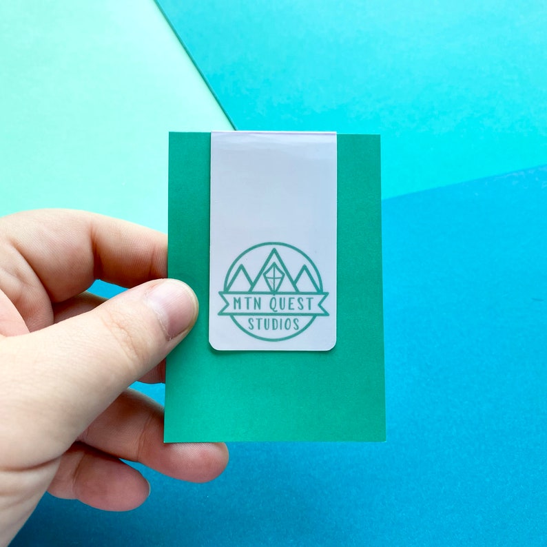 K&ouml;nnte beinhalten: Ein wei&szlig;er B&uuml;roklammer mit einem gr&uuml;nen Logo, auf dem "MTN QUEST STUDIOS" in einem Kreis mit einem Bergketten-Design steht. Die B&uuml;roklammer befindet sich auf einer gr&uuml;nen Karte.