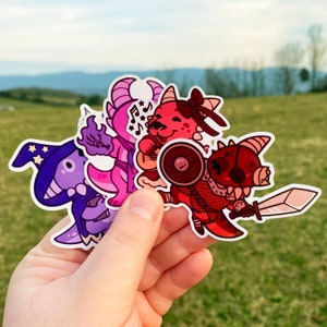 May include: A set of four colorful cartoon dragon stickers. The stickers feature a purple dragon wearing a wizard hat, a pink dragon with wings, a red dragon with a shield, and a red dragon holding a sword.