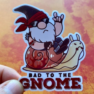 May include: A sticker featuring a cartoon gnome wearing sunglasses and a red bandana, riding a snail with flames on its shell. The text "BAD TO THE GNOME" is printed below the image.