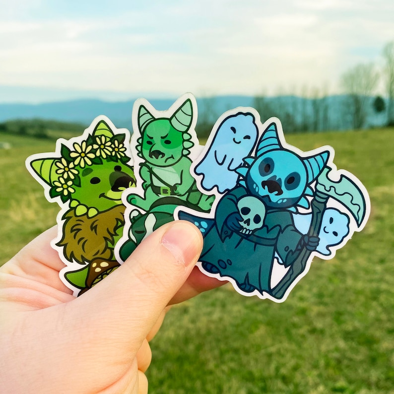 May include: Four colorful cartoon character stickers featuring a green monster, a green monster riding a creature, a blue monster holding a skull, and a white ghost. The stickers are on a hand.