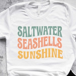 May include: White t-shirt with the words 'SALTWATER SEASHELLS SUNSHINE' in a retro font. The text is in shades of green, coral, and yellow. The shirt is paired with black shorts and a black watch. A clear water bottle is visible on the left.