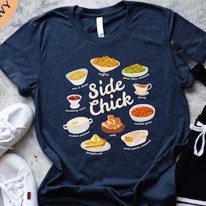 May include: Heather navy blue t-shirt with the words "Side Chick" and illustrations of Thanksgiving side dishes. Dishes include stuffing, green bean casserole, cranberry sauce, mashed potatoes, rolls, and sweet potato casserole.