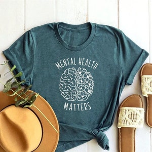 Op de afbeelding: Tealblauw T-shirt met witte tekst die "Mental Health Matters" zegt en een witte omtrek van een brein met bloemen erin.