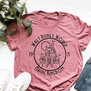 K&ouml;nnte beinhalten: Ein pinkfarbenes T-Shirt mit einer schwarzen Grafik von Cinderellas Schloss in einem Kreis mit dem Text "Walt Disney World 1971 Magic Kingdom".