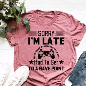 May include: A pink t-shirt with the text "Sorry I'm Late Had To Get To A Save Point" and a black video game controller graphic.