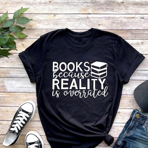 May include: Black t-shirt with white text that reads "BOOKS because REALITY is overrated" and a stack of books graphic. The shirt is laid out on a wooden surface with a pair of black and white sneakers, blue jeans, and a black baseball cap with a white logo.