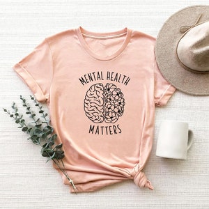 Op de afbeelding: Een roze T-shirt met een zwarte omtrek van een brein met bloemen aan de ene kant. De tekst "Mental Health Matters" is op het T-shirt gedrukt.