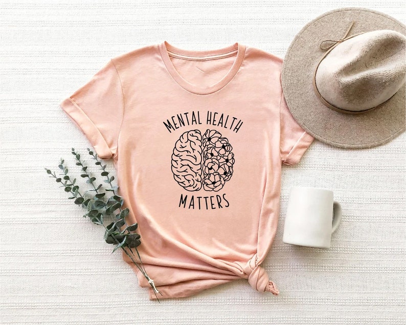 Op de afbeelding: Een roze T-shirt met een zwarte omtrek van een brein met bloemen aan de ene kant. De tekst "Mental Health Matters" is op het T-shirt gedrukt.