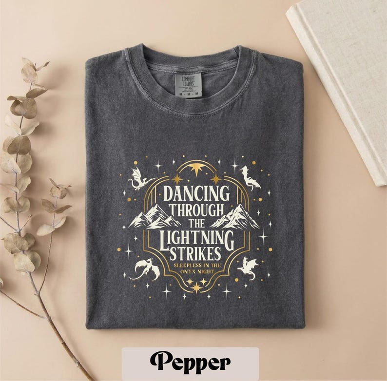 A gray t-shirt with a graphic design featuring a mountain range and stars, along with the text "DANCING THROUGH THE LIGHTNING STRIKES" and "SLEEPLESS IN THE NIGHT".