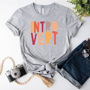 May include: A grey t-shirt with the word "INTROVERT" printed in colourful block letters. The letters are in shades of red, orange, yellow, and pink.