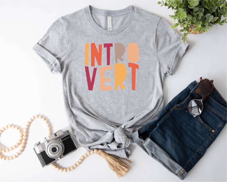 May include: A grey t-shirt with the word "INTROVERT" printed in colourful block letters. The letters are in shades of red, orange, yellow, and pink.