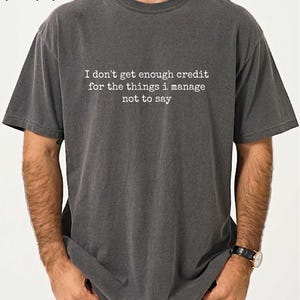 A man wearing a gray t-shirt with the text "I don't get enough credit for the things I manage, not to say" printed on it.