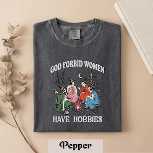 May include: A charcoal gray crew neck t-shirt featuring a graphic of four figures in witch hats sitting around a campfire in a forest. The text reads GOD FORBID WOMEN HAVE HOBBIES. The shirt has a vintage wash finish and a Comfort Colors brand label.