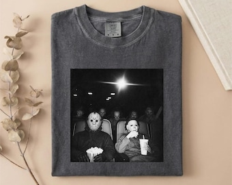 May include: A charcoal gray short-sleeve t-shirt featuring a black and white graphic print of two horror movie characters sitting in a movie theater, one wearing a hockey mask and holding popcorn, the other wearing a white mask and holding a drink.