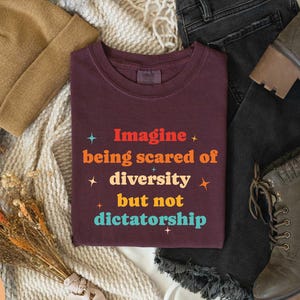 May include: A berry-colored t-shirt with the text "Imagine being scared of diversity but not dictatorship" in colorful lettering. The shirt is paired with a tan beanie, black jeans, and olive-green boots.