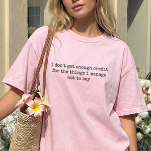 A young woman wearing a pink t-shirt with text on it, standing in front of a building with flowers in the background.