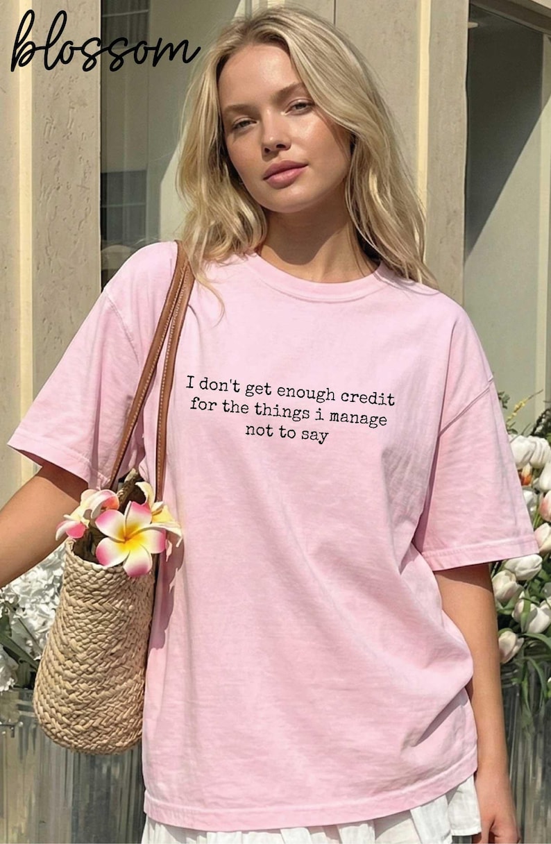 A young woman wearing a pink t-shirt with text on it, standing in front of a building with flowers in the background.