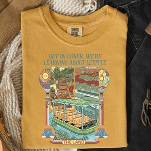 a yellow t-shirt with a colorful illustration of a boat and a sign that says "GET IN LOSER WE'RE LEARNING ABOUT LITTUCE" printed on it.
