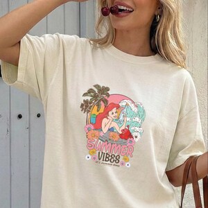 A woman wearing a white t-shirt with a graphic design of a mermaid and the text "Summer Vibes" is smiling and holding a flower.
