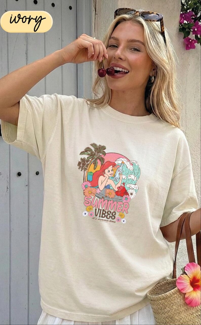 A woman wearing a white t-shirt with a graphic design of a mermaid and the text "Summer Vibes" is smiling and holding a flower.