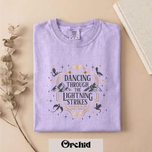 A purple t-shirt with a graphic design featuring a dragon and stars, along with the text "DANCING THROUGH THE LIGHTNING STRIKES" and "SLEEP IN THE WILD ON NIGHT".