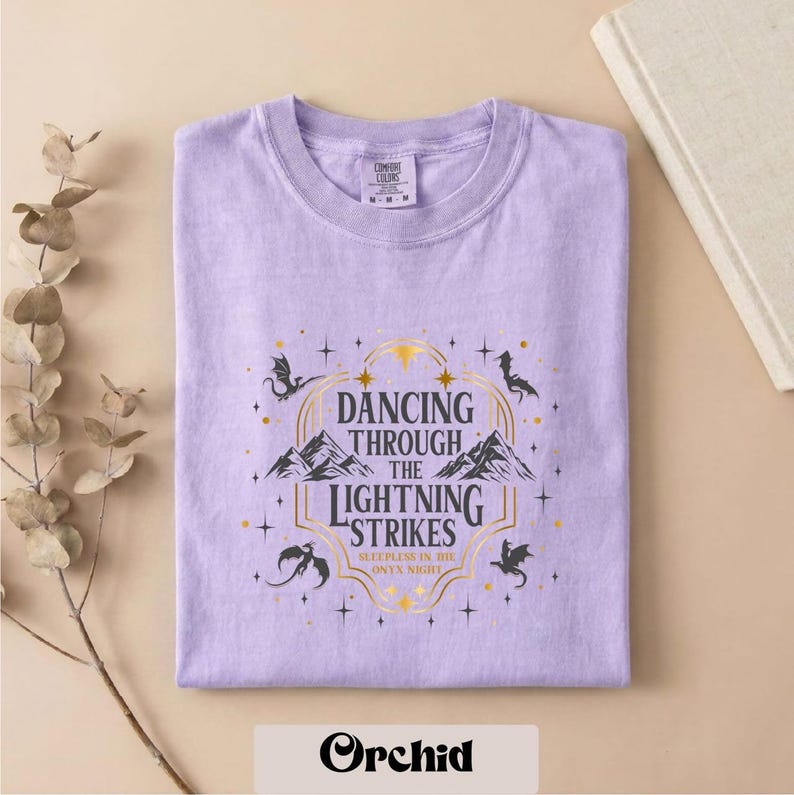 A purple t-shirt with a graphic design featuring a dragon and stars, along with the text "DANCING THROUGH THE LIGHTNING STRIKES" and "SLEEP IN THE WILD ON NIGHT".