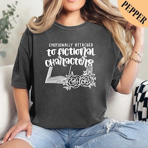 May include: A dark gray t-shirt with the text "Emotionally attached to fictional characters" in white. The text is surrounded by a stylized image of an open book with flowers.