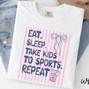 May include: White t-shirt with the text "EAT. SLEEP. TAKE KIDS TO SPORTS. REPEAT" in blue, set against a pink striped background. A bow and a drink graphic are also featured. The word "white" is written in cursive.