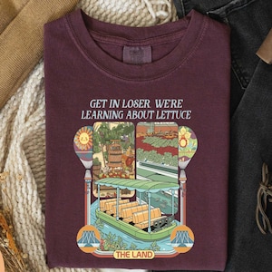 a maroon t-shirt with a colorful graphic design depicting a boat and a garden, along with the text "GET IN LOSER WE'RE LEARNING ABOUT LITTUCE" printed on it.