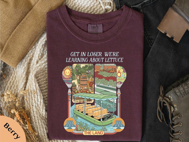 a maroon t-shirt with a colorful graphic design depicting a boat and a garden, along with the text "GET IN LOSER WE'RE LEARNING ABOUT LITTUCE" printed on it.
