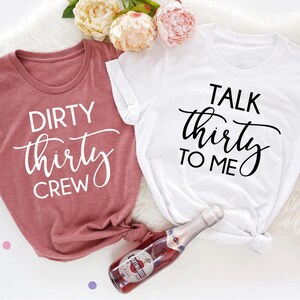 May include: Two t-shirts with a birthday theme. One shirt is pink and says "Dirty Thirty Crew" in white text. The other shirt is white and says "Talk Thirty To Me" in black text.