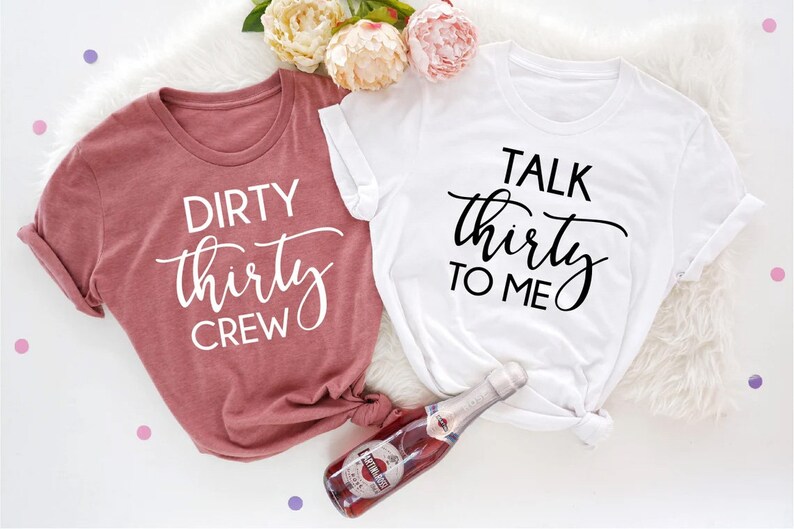 May include: Two t-shirts with a birthday theme. One shirt is pink and says "Dirty Thirty Crew" in white text. The other shirt is white and says "Talk Thirty To Me" in black text.
