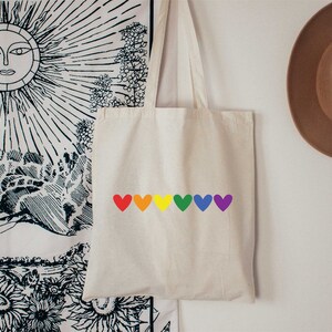 May include: A white tote bag with a rainbow of hearts printed on the front. The hearts are red, orange, yellow, green, blue, and purple.