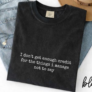 a black t-shirt with white text that reads "I don't get enough credit for the things I manage not to say". The t-shirt is placed on top of a pair of blue jeans and a brown hat, creating a casual and laid-back outfit.