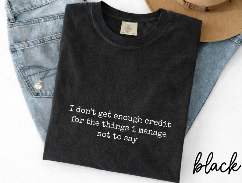 a black t-shirt with white text that reads "I don't get enough credit for the things I manage not to say". The t-shirt is placed on top of a pair of blue jeans and a brown hat, creating a casual and laid-back outfit.