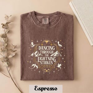A brown t-shirt with a graphic design featuring a mountain range and stars, along with the text "DANCING THROUGH THE LIGHTNING STRIKES ONLY AT NIGHT" printed on it.