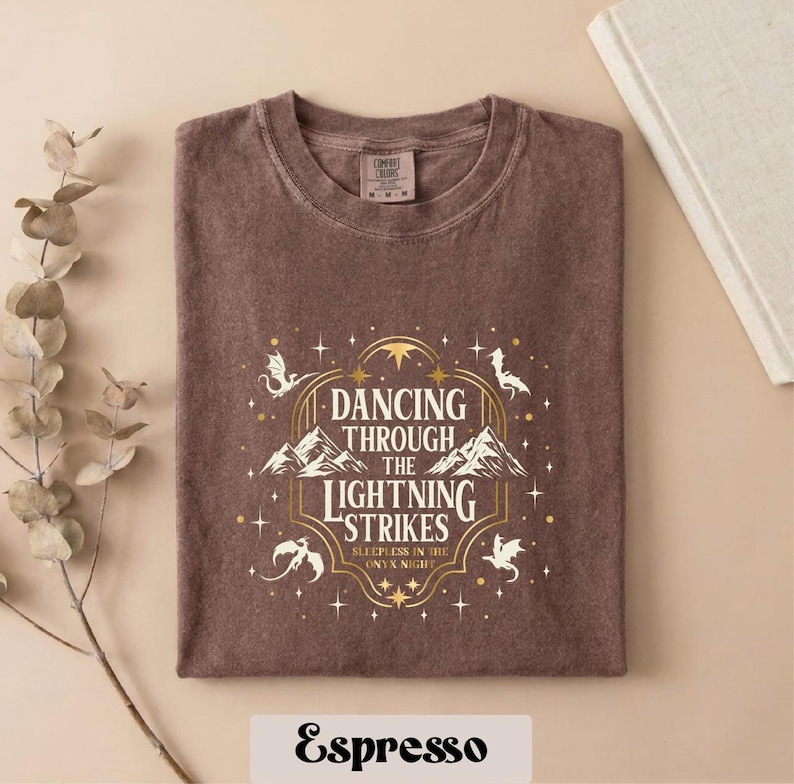 A brown t-shirt with a graphic design featuring a mountain range and stars, along with the text "DANCING THROUGH THE LIGHTNING STRIKES ONLY AT NIGHT" printed on it.