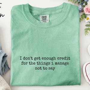 a green t-shirt with a humorous text printed on it, along with a pair of jeans, sneakers, and a floral arrangement.