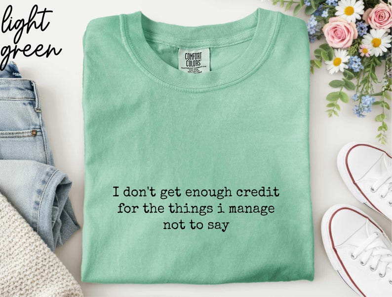 a green t-shirt with a humorous text printed on it, along with a pair of jeans, sneakers, and a floral arrangement.