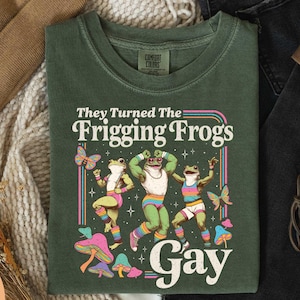 May include: Moss green t-shirt with the text "They Turned The Frigging Frogs Gay" and a graphic of three dancing frogs, butterflies, and mushrooms. The shirt is made of a soft, comfortable material.