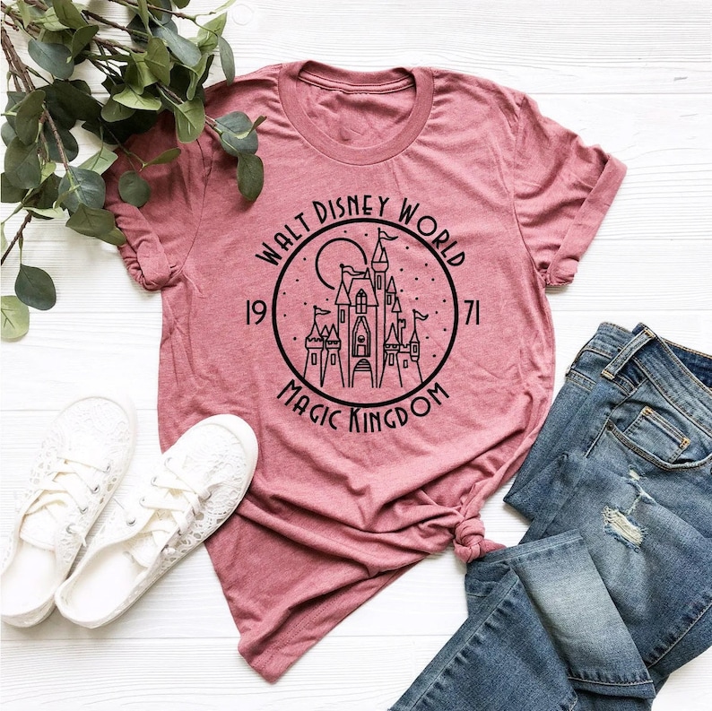 K&ouml;nnte beinhalten: Ein pinkfarbenes T-Shirt mit einer schwarzen Grafik von Cinderellas Schloss in einem Kreis mit dem Text "Walt Disney World 1971 Magic Kingdom".