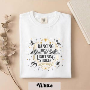 A white t-shirt with a graphic design featuring a dragon and stars, along with the text "DANCING THROUGH THE LIGHTNING STRIKES" and "SLEEP IN THE ONLY NIGHT".