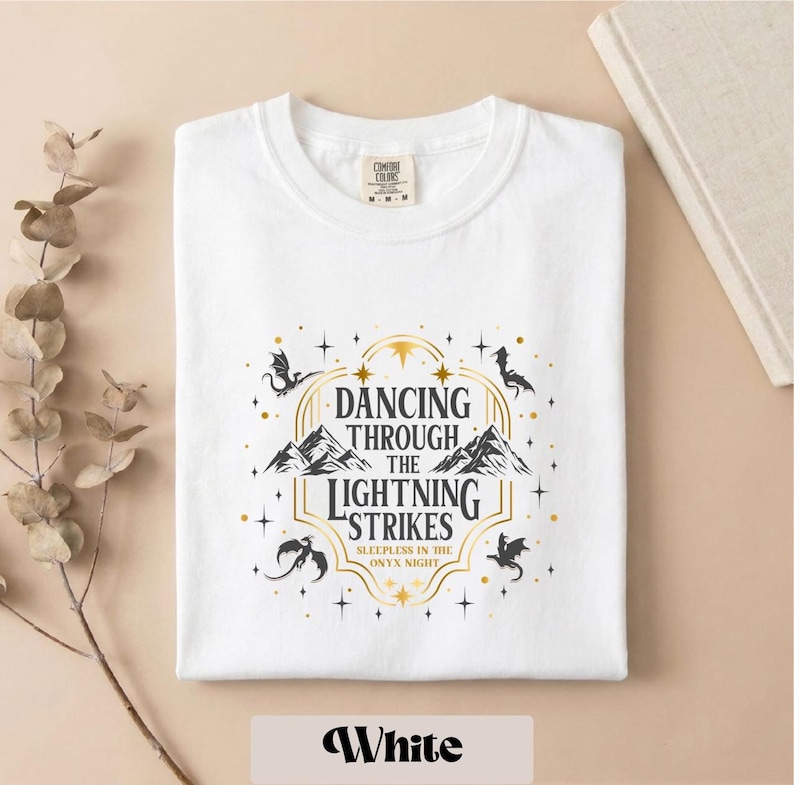 A white t-shirt with a graphic design featuring a dragon and stars, along with the text "DANCING THROUGH THE LIGHTNING STRIKES" and "SLEEP IN THE ONLY NIGHT".